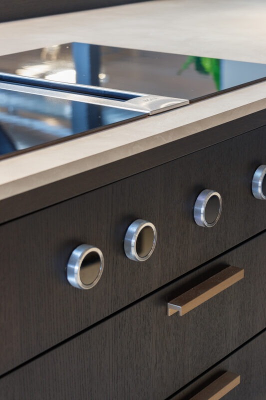 Een detailopname van een luxe, donker eikenhouten keuken met een strak, licht aanrechtblad. In het blad is een inductiekookplaat met geïntegreerde afzuiging verwerkt. Op het donkere frontpaneel direct onder de kookplaat zitten robuuste, roestvrijstalen bedieningsknoppen met een kartelrand en donkere binnenkant. Daaronder bevinden zich lades met strakke, bronskleurige handgrepen.