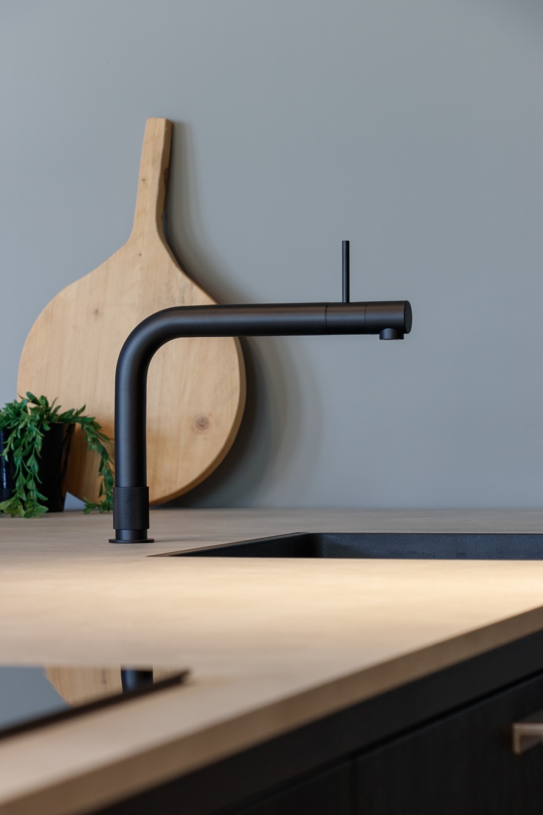 Een detailopname van een luxe, matzwarte keukenkraan met een minimalistisch design en een subtiele kartelstructuur op de voet. De kraan is gemonteerd op een licht, modern aanrechtblad naast een zwarte spoelbak. Op de achtergrond leunt een grote, ronde houten serveerplank tegen een strakke, grijze wand.