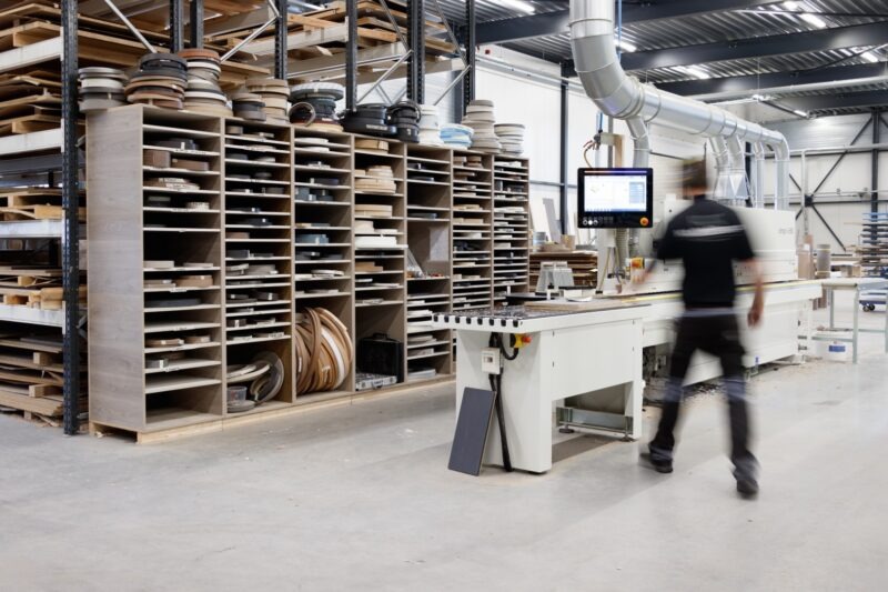 Een overzichtsfoto van de moderne werkplaats van Klomp Keukens. Een grote, open kast staat vol met talloze rollen gesorteerd kantfineer of kantbanden in verschillende kleuren. Rechts staat een geavanceerde computergestuurde Olimpic kantenaanlijmmachine met een touch-screen controller. Een bewogen persoon loopt langs de machine en suggereert dagelijkse activiteit. Op de achtergrond hoge stellingen met plaatmateriaal en industriële luchtzuiginstallaties.