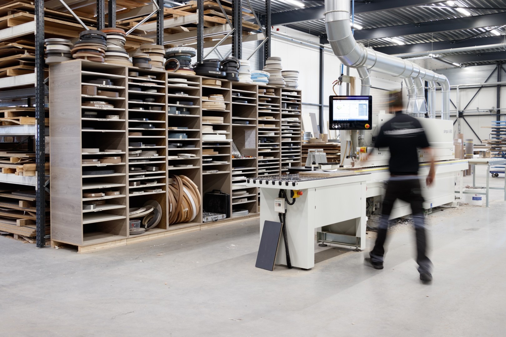 Een overzichtsfoto van de moderne werkplaats van Klomp Keukens. Een grote, open kast staat vol met talloze rollen gesorteerd kantfineer of kantbanden in verschillende kleuren. Rechts staat een geavanceerde computergestuurde Olimpic kantenaanlijmmachine met een touch-screen controller. Een bewogen persoon loopt langs de machine en suggereert dagelijkse activiteit. Op de achtergrond hoge stellingen met plaatmateriaal en industriële luchtzuiginstallaties.